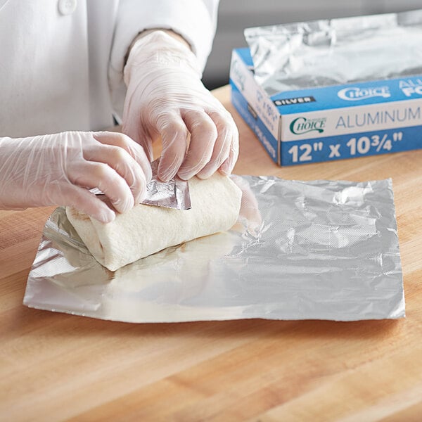 A person in gloves is wrapping food in a Choice blue and white box of interfolded foil sheets.