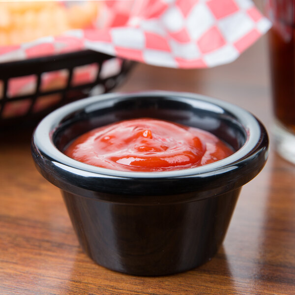 A Tablecraft black melamine ramekin filled with ketchup on a table with a basket of fries.