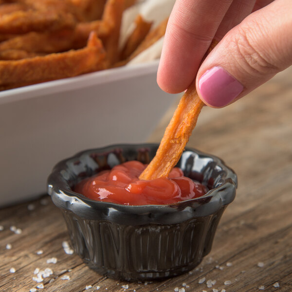 A person using a Carlisle black scalloped ramekin to dip a french fry into ketchup.