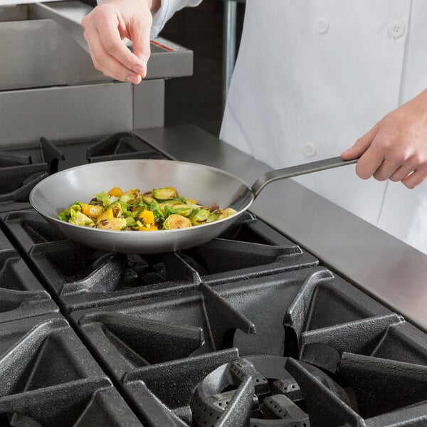 A chef cooking vegetables in a Matfer Bourgeat carbon steel fry pan on a stove.