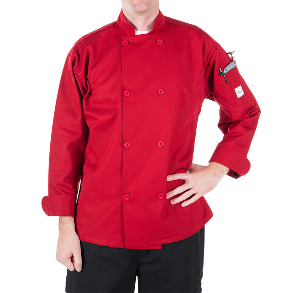 A man standing on a counter in a professional kitchen wearing a red Mercer Culinary chef jacket.