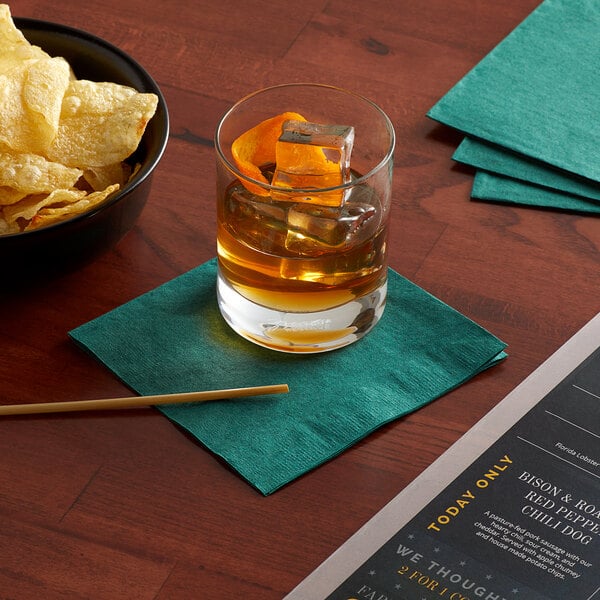 A table with a glass of liquid and a bowl of potato chips with a Choice Hunter Green beverage napkin.