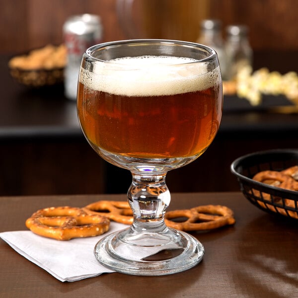 An Anchor Hocking schooner glass filled with beer sitting on a table with a basket of pretzels.
