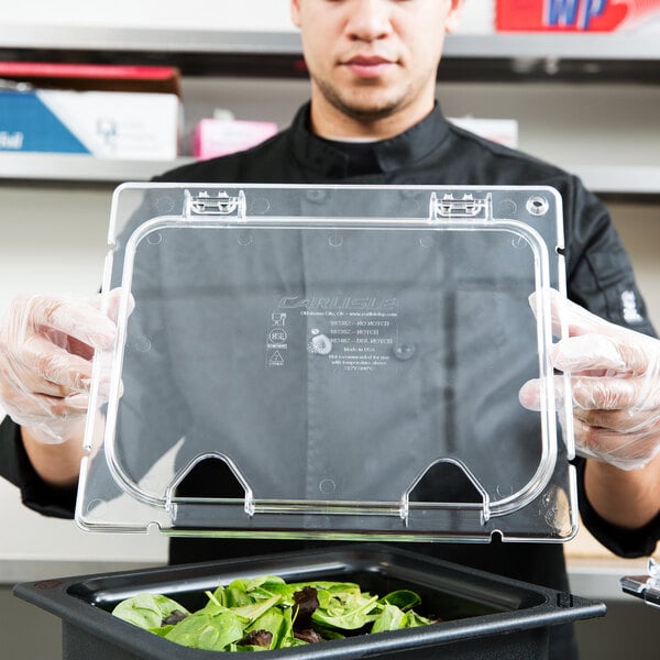A man in a chef's uniform holding a Carlisle clear plastic food pan lid over a clear plastic container on a counter.