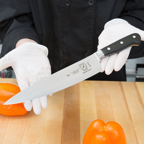 A person in gloves holding a Mercer Renaissance forged riveted chef's knife on a table in a professional kitchen.