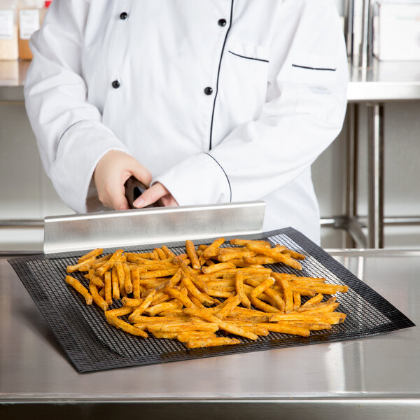 A person in a white coat using a Baker's Mark mesh screen to cook french fries on a black tray.