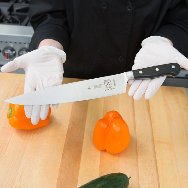 A person in a white glove using a Mercer Culinary Renaissance Forged Chef's Knife to cut a pepper on a wooden cutting board.