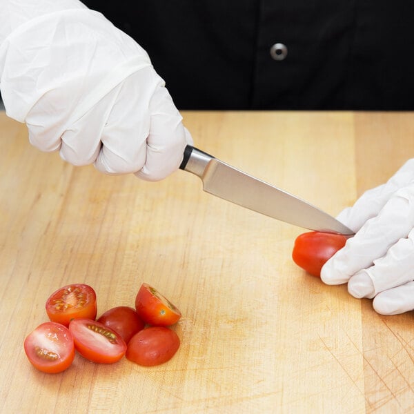 A hand in a white glove uses a Mercer Culinary Renaissance Forged Paring Knife to cut a tomato on a wooden cutting board.