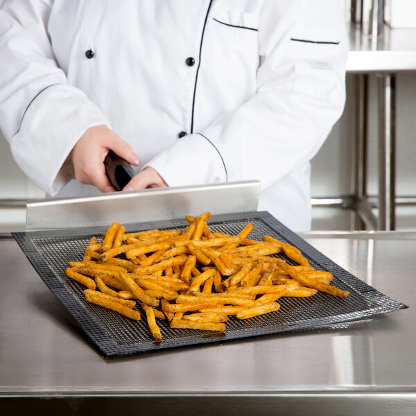 A chef in a white coat using a black Baker's Mark mesh screen to cook french fries on a metal tray.