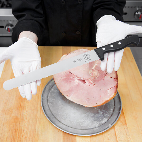 A hand holding a Mercer Culinary Renaissance Slicer with a serrated edge over a piece of ham.