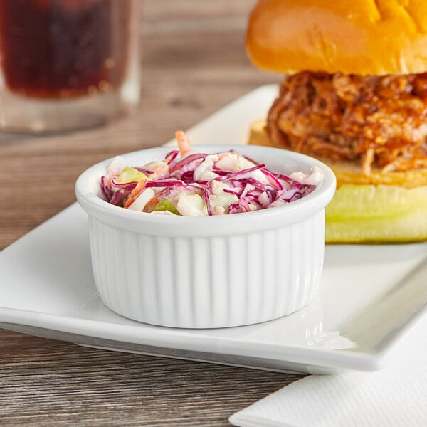 A plate with a sandwich and a bowl of coleslaw served on a table with an Acopa Bright White Fluted Porcelain Ramekin filled with coleslaw.