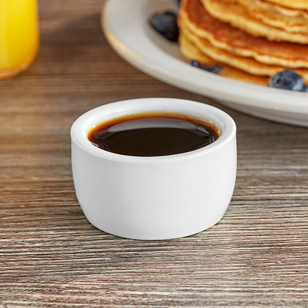 A white cup of syrup on a table next to a plate of pancakes.
