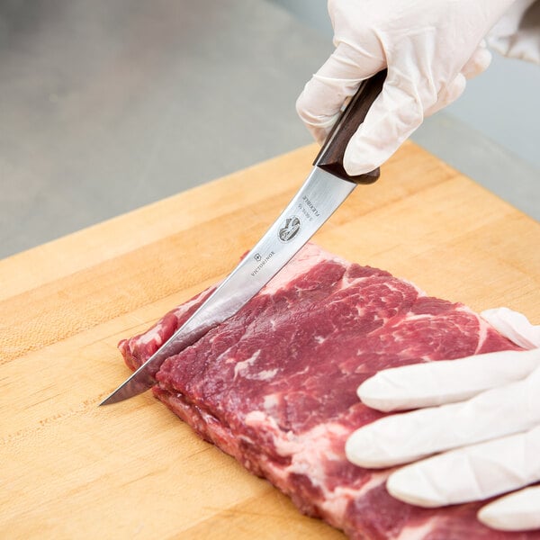 A person wearing a glove with a wooden-handled Victorinox curved boning knife cutting meat on a counter.