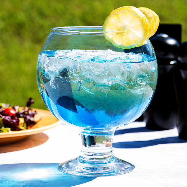 A Libbey Super Globe Fish Bowl Glass with blue liquid and lemon slices on a white table.