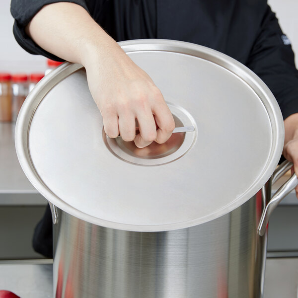 A woman in a black chef's uniform holding a large silver Vollrath pot with a stainless steel lid.