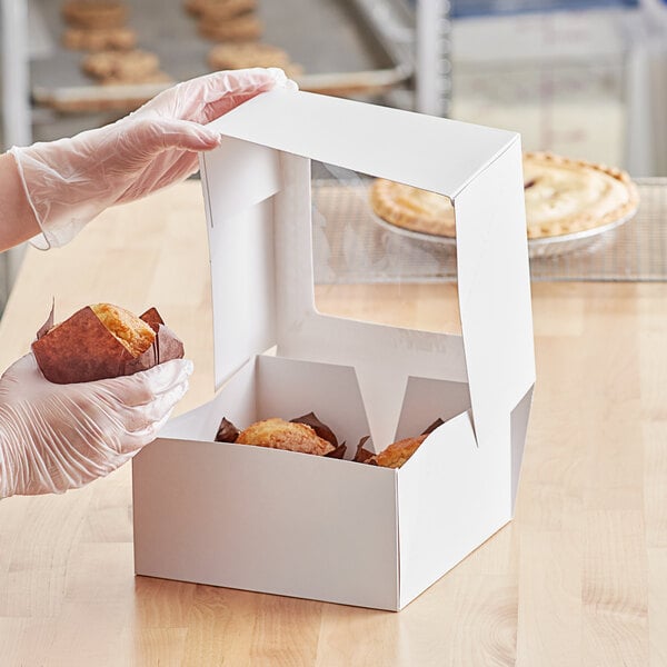 A person in gloves holding a white Auto-Popup window bakery box full of muffins.