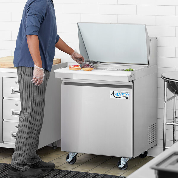 An Avantco stainless steel refrigerated sandwich prep table on a counter in a professional kitchen with a man wearing gloves.