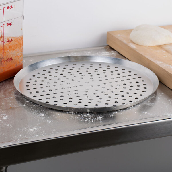 An American Metalcraft 18" heavy weight aluminum perforated pizza pan on a counter with a dough ball on it.