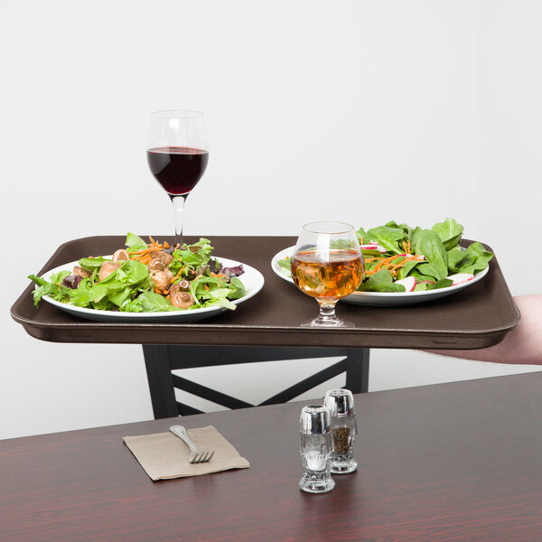 A person holding a Carlisle brown non-skid tray with a plate of salad and a glass of brown liquid.