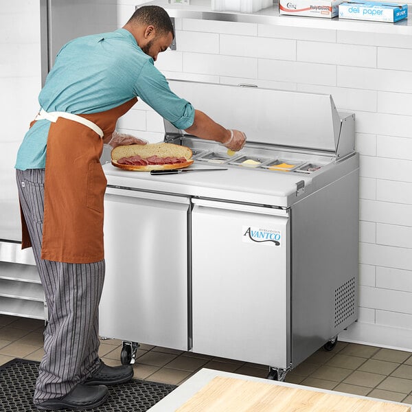 An man in an apron using an Avantco stainless steel sandwich prep table to make sandwiches.