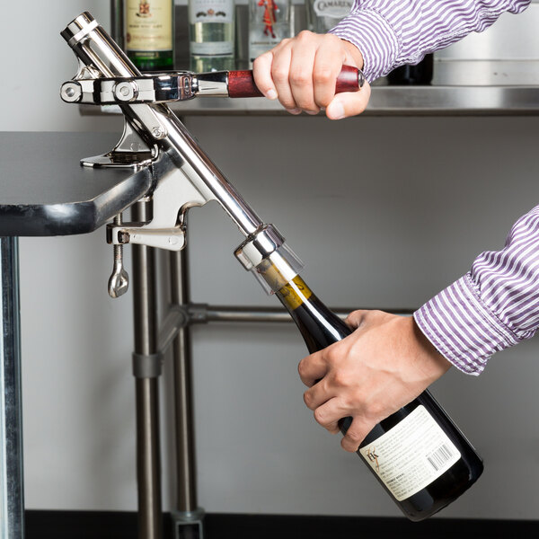 A person using a Franmara Cedon nickel-plated counter mount wine bottle opener to open a bottle of wine on a counter in a bar.