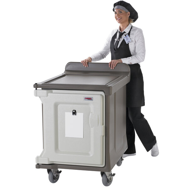 A woman in a chef hat and apron pushing a Cambro meal delivery cart with a large grey container inside.