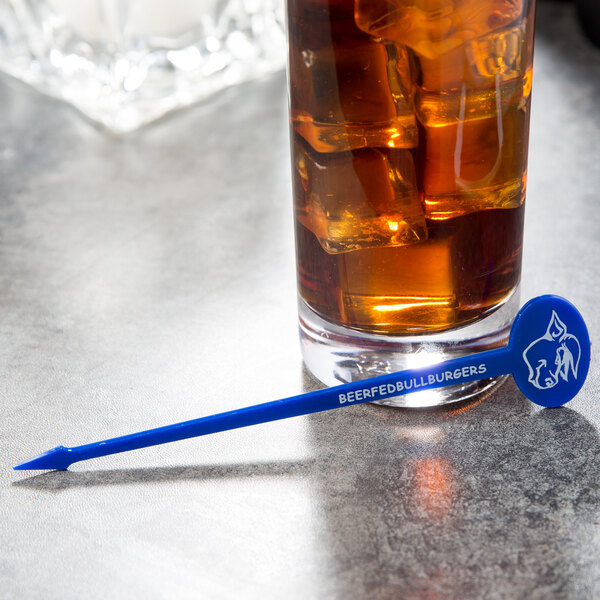 A glass of liquid with ice and a blue oval stirrer with a blue toothpick sitting on top.