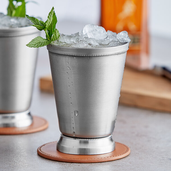Two Acopa stainless steel mint julep cups with ice and mint leaves on a coaster.
