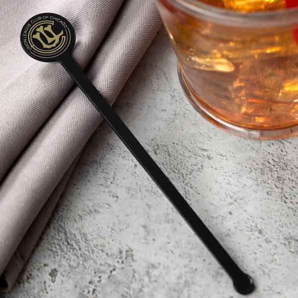 A WNA Comet black drink stirrer with a gold logo on a napkin next to a glass of brown liquid.