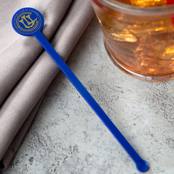 A glass of brown liquid with a blue WNA Comet Spirit Customizable disc stirrer.