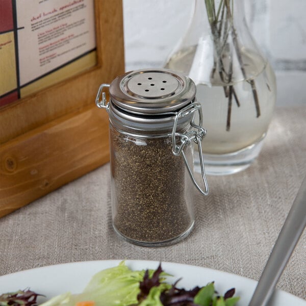 A Tablecraft glass jar with a stainless steel clip-top lid used as a pepper shaker on a table.