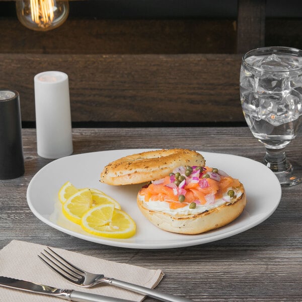 A Libbey bright white porcelain coupe plate with a bagel and salmon on a table with a fork and lemon slices.