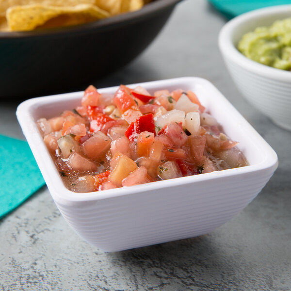 A white square ribbed melamine ramekin filled with salsa on a table next to a bowl of guacamole.