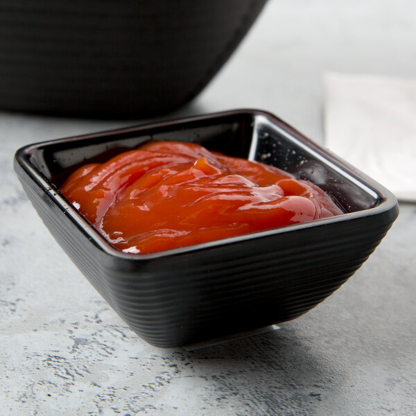 A black Tablecraft square ribbed melamine ramekin filled with red sauce on a table.