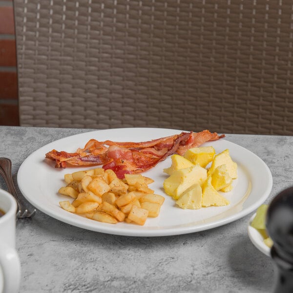 A close up of a Libbey Porcelana narrow rim porcelain plate with bacon, eggs, and potatoes on it on a table.