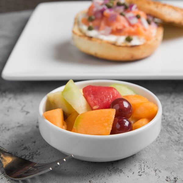 A bowl of fruit in a white Libbey porcelain bowl on a table with a bagel and a fork.