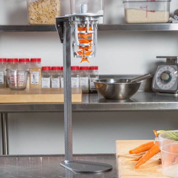 A Tellier manual upright carrot peeler on a counter with carrots in a clear plastic container.