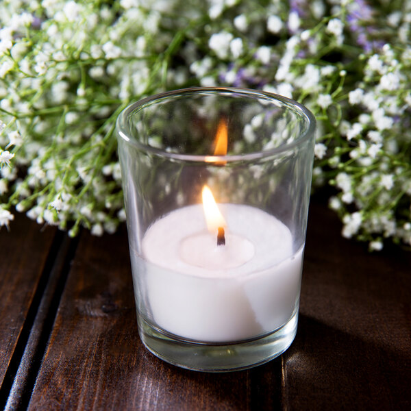 A Leola clear glass wax filled votive candle on a wooden table in a room with white flowers.