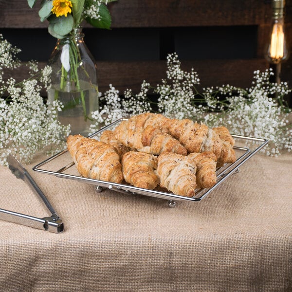 A Clipper Mill chrome plated iron square wire basket with croissants on a table.