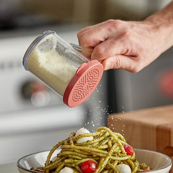 A hand holding a Cambro polycarbonate shaker with a pink lid sprinkling cheese over a bowl of pasta.