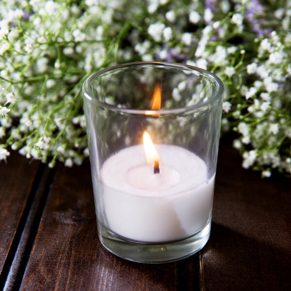 A Leola clear glass votive candle on a wooden table in a room with white flowers.