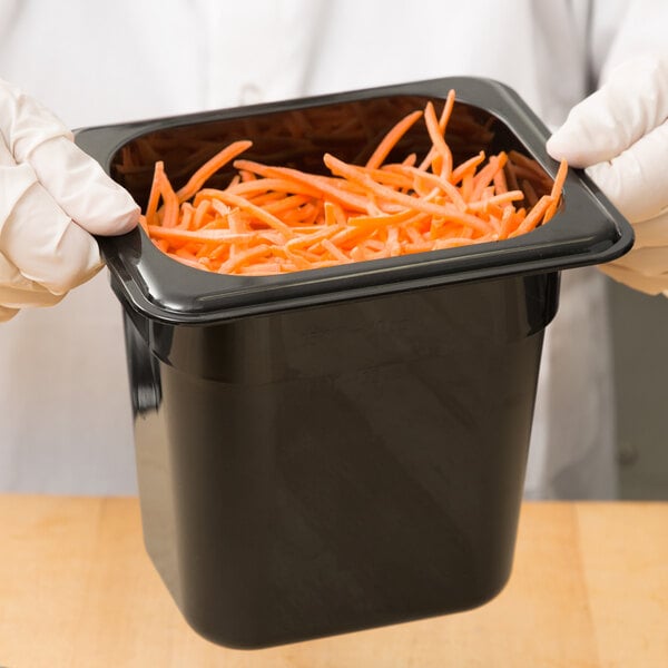 A person in gloves holding a black Cambro plastic food pan full of carrots.