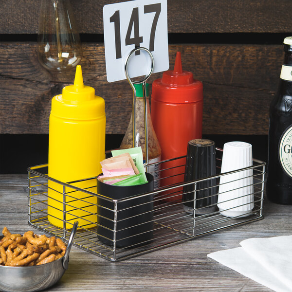 A Clipper Mill stainless steel rectangular wire caddy holding condiments and a bottle of beer on a table.