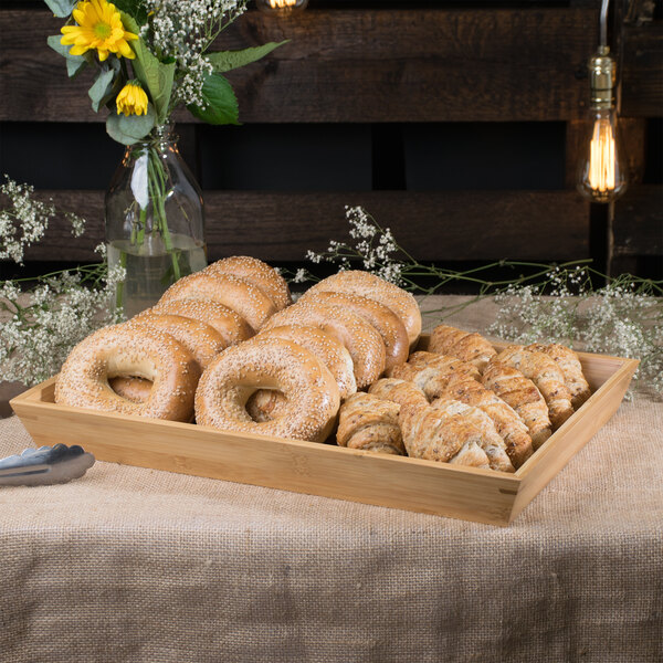 A Clipper Mill bamboo tray filled with bagels and pastries on a table in a bakery display with flowers.