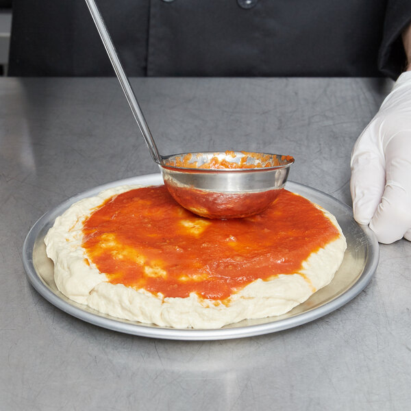 A person using a ladle to put sauce on a pizza on a pizza pan.