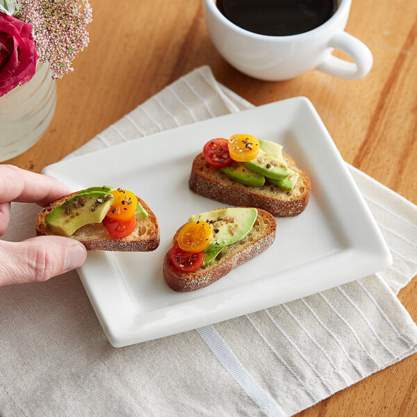 A hand holding a Tuxton white rectangular plate with a slice of bread topped with avocado and tomatoes.