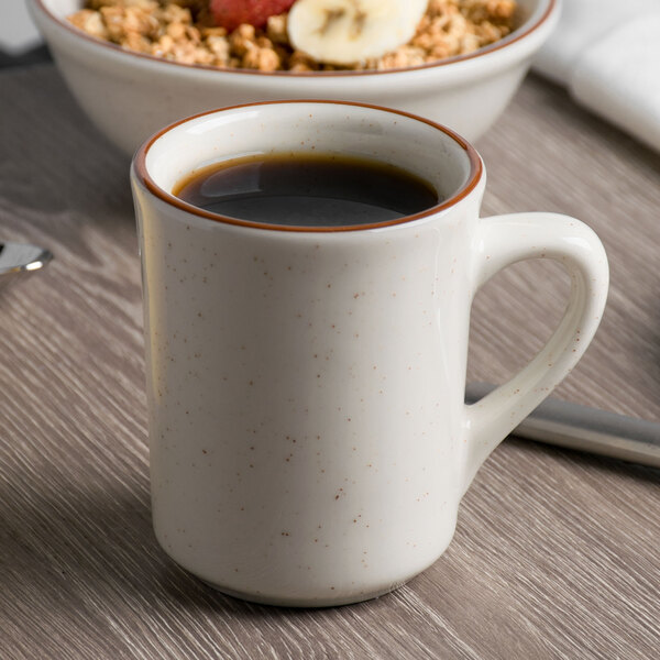 A cup of coffee in a Tuxton brown speckle mug on a table with a bowl of cereal and fruit.
