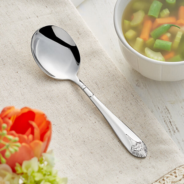 An Acopa Monaca stainless steel bouillon spoon with a flower on it on a table next to a bowl of soup.
