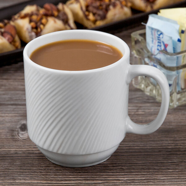 A Libbey bright white stacking mug filled with brown liquid sits on a table next to a tray of pastries.