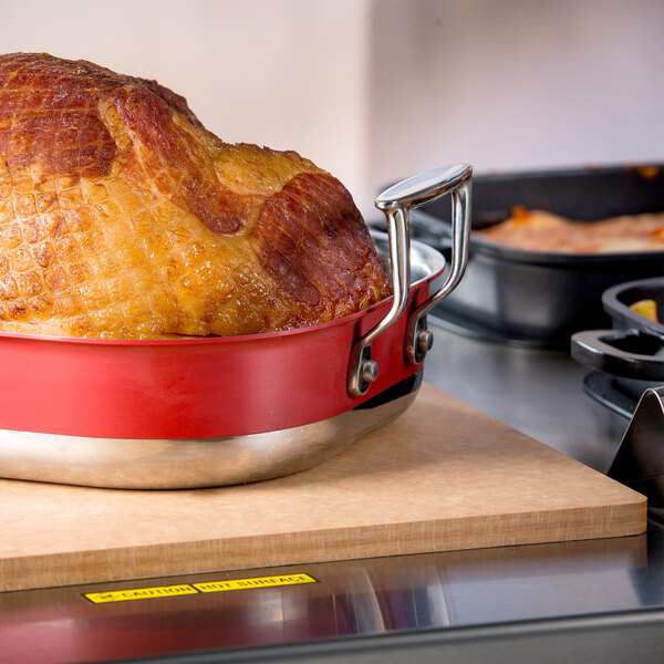 A cooked ham in a red pan on a Tablecraft natural solid double well cutting board.